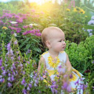 Bild på en bebis som sitter ute i naturen omringad av växter och blommor med solljus bakom sig.