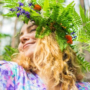 En flicka som bär en blomsterkrans på huvudet med gröna blad och färgglada blommor. Hon har långt, ljust hår och ler mjukt. hon har ögonen slutna medan sommar solen skiner.