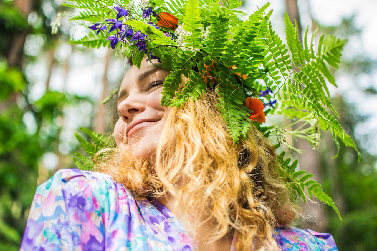 En flicka som bär en blomsterkrans på huvudet med gröna blad och färgglada blommor. Hon har långt, ljust hår och ler mjukt. hon har ögonen slutna medan sommar solen skiner.