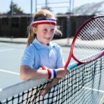Barn med tenniskläder och tennisracket som står vid ett tennisnät utomhus.