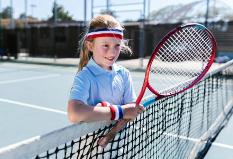 Barn med tenniskläder och tennisracket som står vid ett tennisnät utomhus.