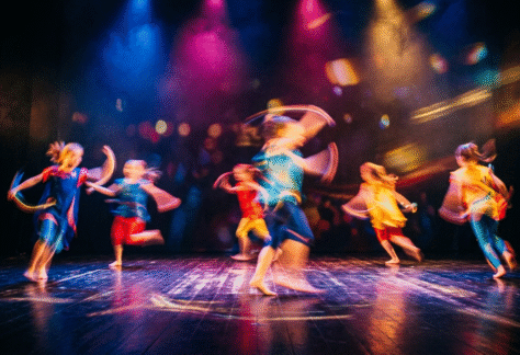 Bild på barn som dansar på en scen. Lampor i blå, lila, gul lyser på scenen.