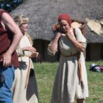 Tre personer med vikingakläder, en man och två kvinnor. Ena kvinnan bär på en korg som hon håller med båda händerna vid axeln. Bakgrunden består av gräs på marken med ett medeltids hus