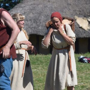 Tre personer med vikingakläder, en man och två kvinnor. Ena kvinnan bär på en korg som hon håller med båda händerna vid axeln. Bakgrunden består av gräs på marken med ett medeltids hus