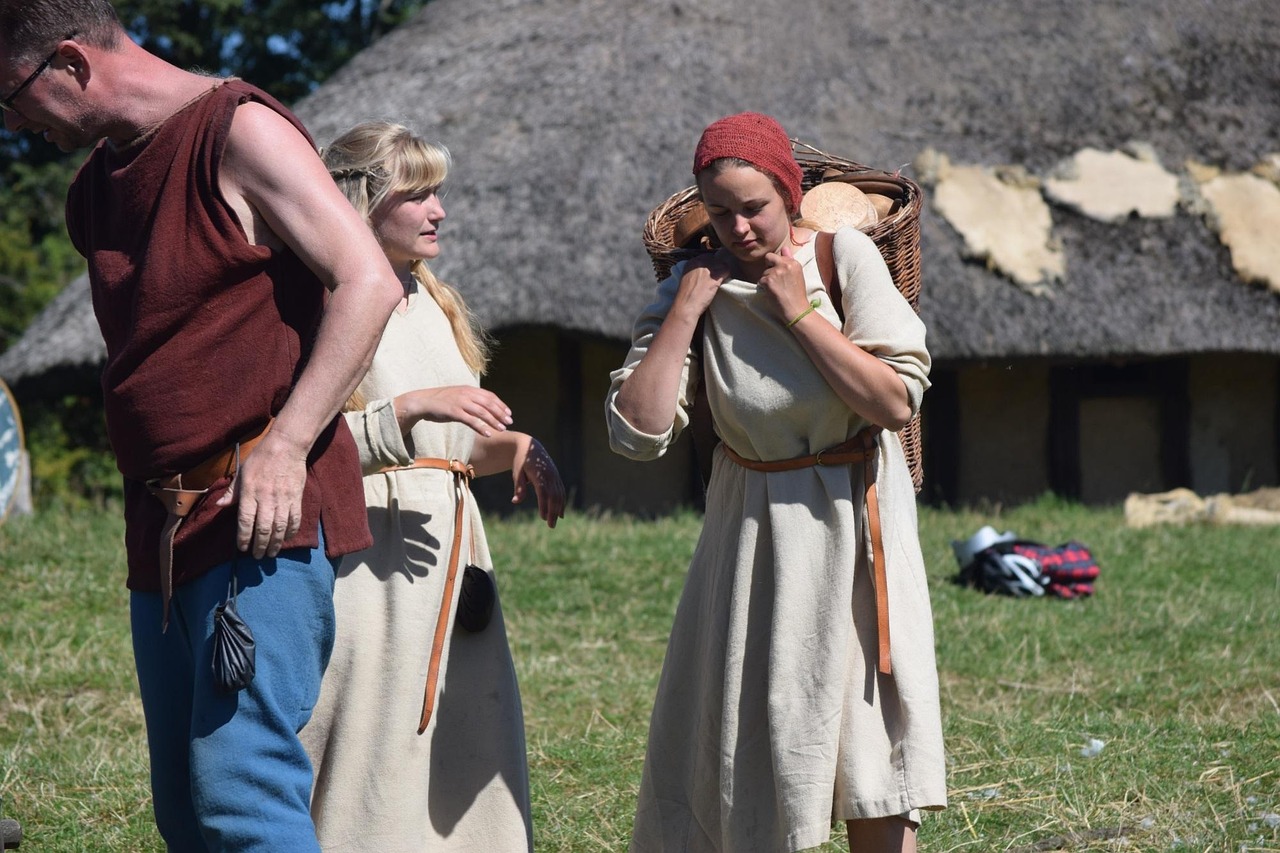 Tre personer med vikingakläder, en man och två kvinnor. Ena kvinnan bär på en korg som hon håller med båda händerna vid axeln. Bakgrunden består av gräs på marken med ett medeltids hus
