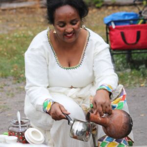 En eritreansk kvinna sitter utomhus och häller upp en dryck. Hon är klädd i en vit traditionell klänning med färgglada detaljer runt halsen och ärmarna.