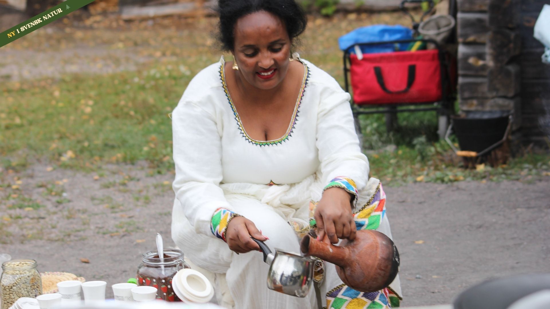 En eritreansk kvinna sitter utomhus och häller upp en dryck. Hon är klädd i en vit traditionell klänning med färgglada detaljer runt halsen och ärmarna.