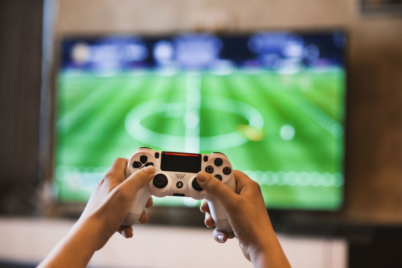 En person som spelar Fifa, man ser fotbollsplanen på TV:n och två händer som håller i en kontroll.