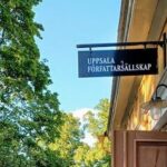 Ingången till ett gult äldre hus på höger sida med en skylt där det står "UPPSALA FÖRFATTARSÄLLSKAP". Lövverk från träd på vänstra sidan.