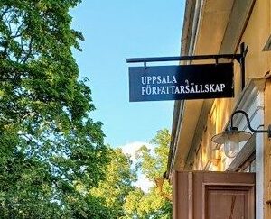 Ingången till ett gult äldre hus på höger sida med en skylt där det står "UPPSALA FÖRFATTARSÄLLSKAP". Lövverk från träd på vänstra sidan.