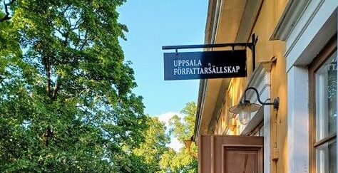 Ingången till ett gult äldre hus på höger sida med en skylt där det står "UPPSALA FÖRFATTARSÄLLSKAP". Lövverk från träd på vänstra sidan.