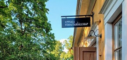 Ingången till ett gult äldre hus på höger sida med en skylt där det står "UPPSALA FÖRFATTARSÄLLSKAP". Lövverk från träd på vänstra sidan.