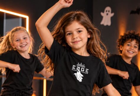 Två flickor och en pojke i svarta T-shirts som dansar i en mörk halloweenutsmyckad danslokal . T-shirten på flickan i mitten har ett vitt tryck där det står "Edge dance studios" och ett spöke.