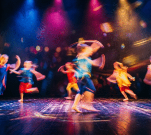 Bild på barn som dansar på en scen. Lampor i blå, lila, gul lyser på scenen.